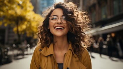 A joyful woman with curly hair and glasses laughing on an urban street with a backdrop of trees with golden leaves.
