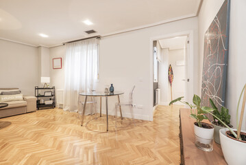 Small living room in a house with circular glass dining table with transparent methacrylate chairs, window with semi-transparent curtains and herringbone French oak parquet floors