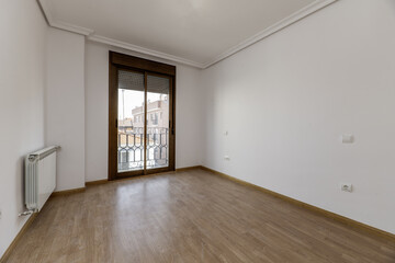 Empty room of a house with wooden floors, plaster moldings on the ceiling, a large dark aluminum and glass window and white aluminum radiator on the wall