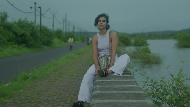 Slow motion shot of a young Indian woman dressed in an elegant purple dress, stands gracefully on the banks of a tranquil river. Her hair flows in the breeze as she gently touches her hair.