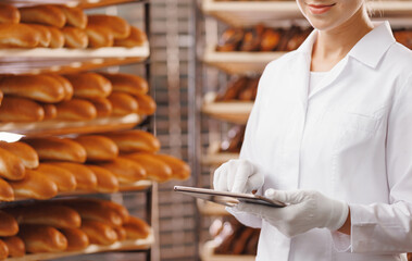 Baker woman in chef uniform use tablet computer for control quality of craft bread in bakery factory, sun light. Banner modern food industry