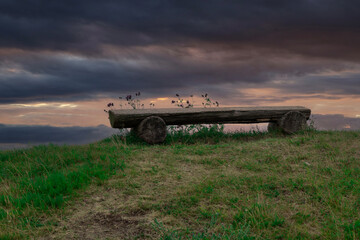 A bench made from a log is placed on top of a hill by the sea. Dramatic sky in the background