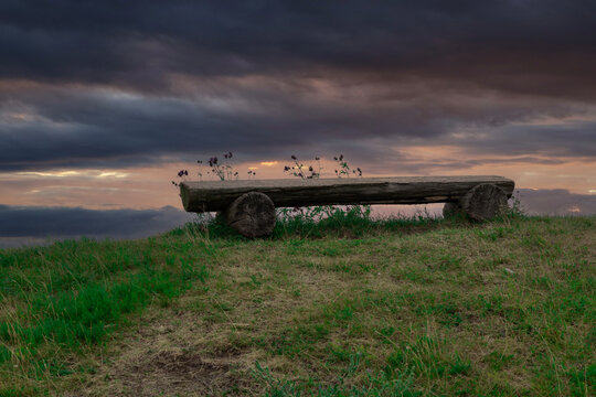 A bench made from a log is placed on top of a hill by the sea. Dramatic sky in the background