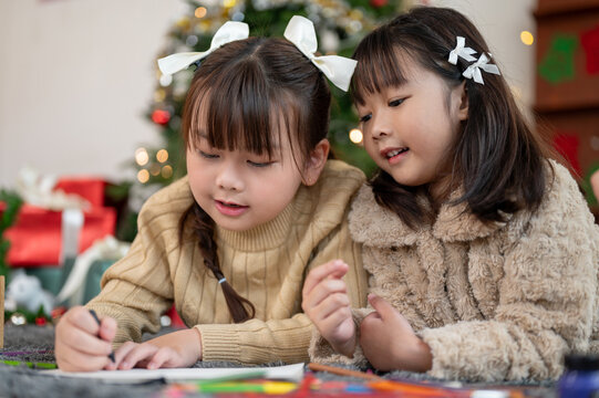 A cute Asian girl is helping her sister making a Christmas card while lying on the floor together.