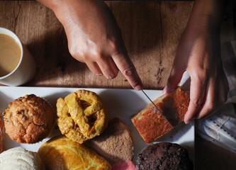 Woman's hand taking a piece of Mexican sweet bread from a white plate on a wooden table. Concept of hands handling food, cutting a Mexican square pudding with a knife, lay flat, top view.