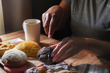Woman's hand taking a piece of Mexican sweet bread from a white plate on a wooden table. Concept of hands handling food, cutting a Mexican chocolate muffin with a knife, side view, horizontal.