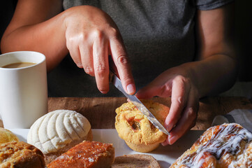 Woman's hand taking a piece of Mexican sweet bread from a white plate on a wooden table. Concept of hands handling food, cutting a pineapple stuffed muffin with a knife, front view, horizontal image.