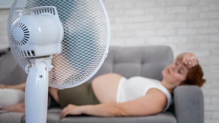 Pregnant woman suffers from heat and lies on the sofa near the electric fan. 