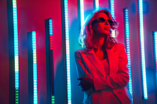 Young beautiful hipster woman in street style fashion concept. Hot model wearing white suit and sunglasses. Female posing in red neon light in studio interior