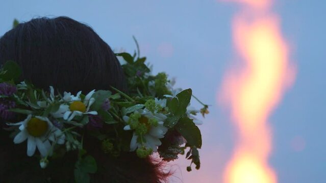 hand held close up shot of woman with a wreath of meadow flowers on her head watching a huge bonfire flame