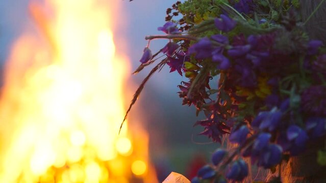 hand held close up shot of woman with a wreath of meadow flowers on her head watching a huge bonfire flame