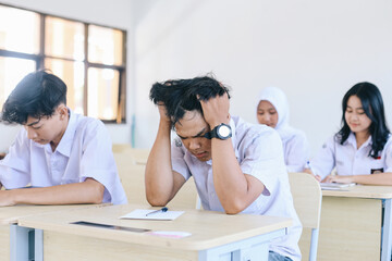 Young Asian male student holding head having headache while sitting at desk during exam in school
