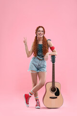 Stylish young hippie woman with guitar showing V-sign on pink background