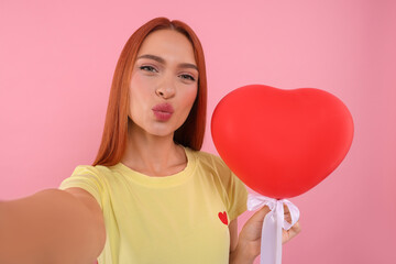 Beautiful woman with red heart shaped balloon taking selfie and sending air kiss on pink background