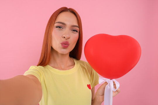 Beautiful woman with red heart shaped balloon taking selfie and sending air kiss on pink background
