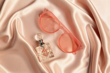 Perfume and pink sunglasses on beige silk fabric, flat lay