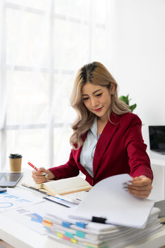 Asian female financial analyst or accountant uses laptop to work on financial audit and budgeting using calculator, checking balance sheet, working with company quarterly report. Vertical image