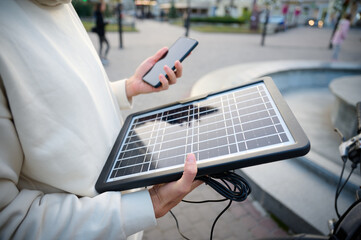 Young pretty woman sitting on the bench and charging her phone with a portable solar panel. Solar...