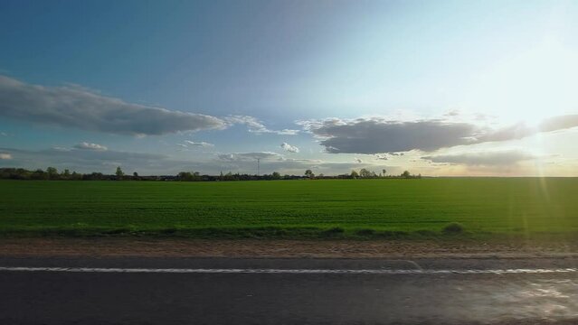 Highway along green meadows and fields in rural area. Background. View through right side window on passenger seat of car van wagon in motion. Roadway out of town. Sunny weather, evening