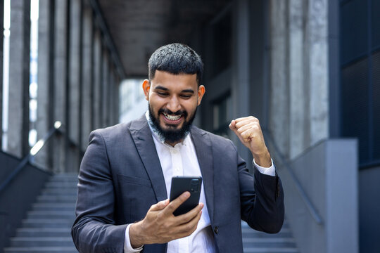 Close-up photo of a young arab man businessman standing on the street near the office and looking at the phone, happy with good news, reading a message, showing a victory yes gesture with his hand
