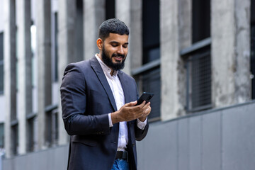 A young Muslim male businessman in a business suit walks down the street near office buildings and uses the phone, calls, reads the news, texts