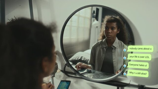 Black teen girl sitting in front of mirror in bedroom, receiving bullying phone messages from Internet haters, feeling stress and looking at reflection. CG text popping up on side