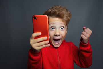 caucasian boy in red jacket looks at his red smartphone with a very surprised expression in a studio on a gray background. - obrazy, fototapety, plakaty