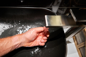 A man's hand touches the water and washes his hands with soap. A black sink with a silver faucet next to an oval mirror and a shelf with hand paper towels. Close-up 