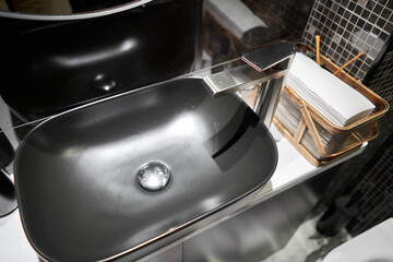 A black sink with a silver faucet next to an oval mirror and a shelf with hand paper towels. Close-up of an elegant faucet in the bathroom sink next to stylish decorations.