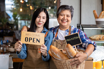 Happy Asian senior elderly retired woman breads cafe coffee small business owner looking at camera with young daughter wearing apron holds open signboard and promotion sign standing smiling together - Powered by Adobe