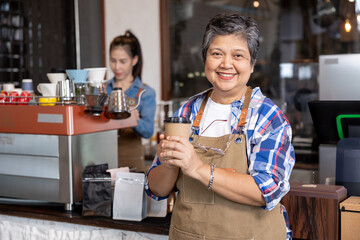 A gray-haired retired Asian businesswoman stands in front holding a brown disposable coffee cup, smiling brightly. There was an employee standing in front taking photos promoting the small family coff - Powered by Adobe