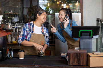 Retired single mother business woman grinds coffee beans smiles Asian daughter coffee maker counter daughter answering phone, taking orders front cash register giving her mother thumbs up. small cafe - Powered by Adobe