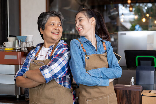 Two Asian Baristas Or Coffee Makers, Mother And Daughter Standing With Arms Crossed Or Confident Poses Looking At Each Other And Smiling In A Coffee Shop. Happy Working With Small Businesses Together.