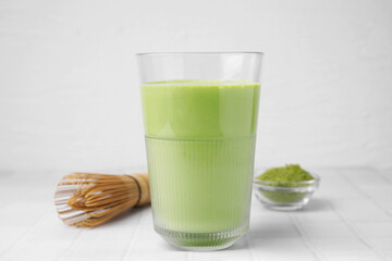 Glass of tasty matcha smoothie, powder and bamboo whisk on white tiled table, closeup