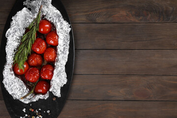 Aluminum foil with delicious baked tomatoes and rosemary on wooden table, top view. Space for text