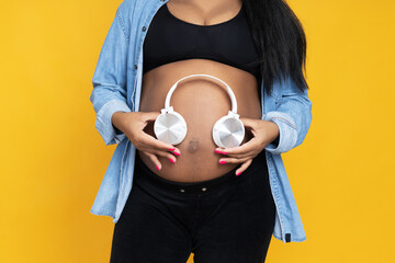 Pregnant African American woman with headphones on yellow background.