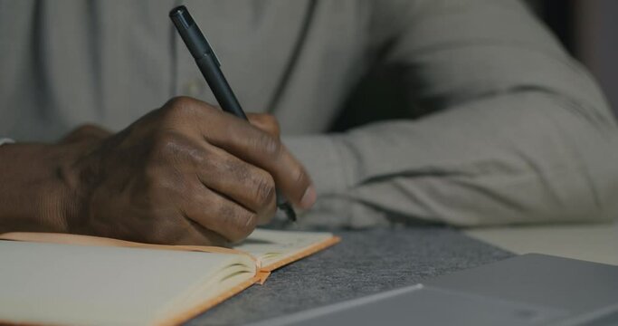 Close-up of male hands writing information in notebook while African American man working at desk at night. Business and job concept.