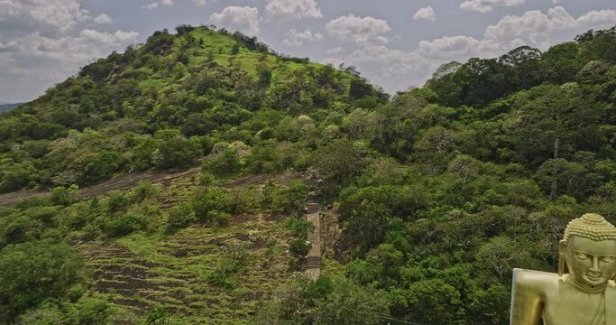 Dambulla Sri Lanka Aerial v2 low drone flyover Uyanwaththa Royal Buddhist Temple capturing golden Buddha on the hillside towards the Jungle Isigili Kanda mountain - Shot with Mavic 3 Cine - April 2023