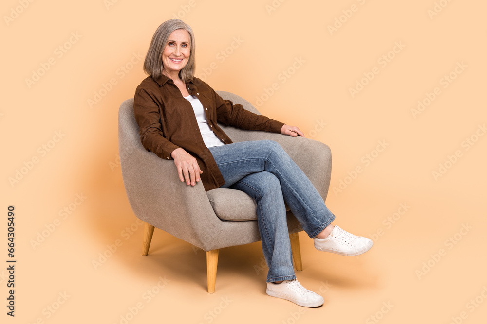 Sticker Full body photo of cheerful senior woman sitting soft gray comfy chair waiting room new furniture shop isolated on beige color background