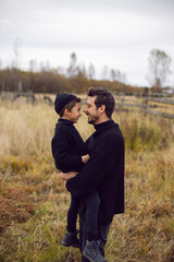 portrait stylish father and son in black clothes stand in a field with an old wooden horse fence in the fall. the man holds his son in his arms in front of him and smiles