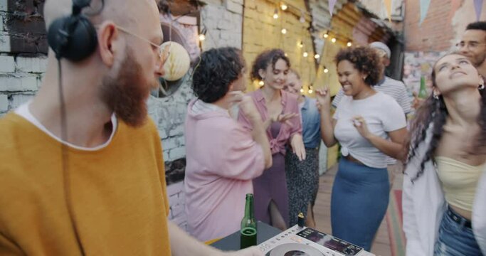 Professional DJ mixing music at open air party while group of young people dancing having fun. Youth culture and entertainment concept.