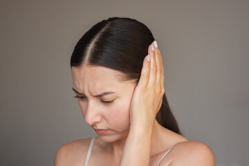 Fototapeta premium a young brunette woman is experiencing acute pain in her ear. She frowns, closes her eyes, presses her ear with her hand. Suffers from otitis.