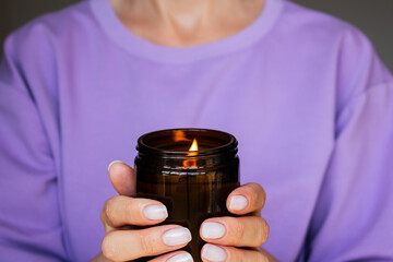 close-up of a dark brown glass jar with a burning candle in the hands of a woman in a purple T-shirt