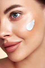 Beautiful woman applying moisturizer cream on her face. Photo of smiling woman with perfect makeup on beige background. Beauty concept