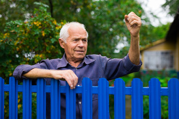 Old elderly farmer stands at fence of his country house and threatens someone with his fist