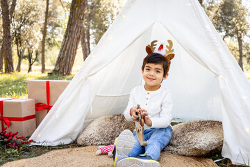 Smiling child boy playing with Christmas decoration in outdoor teepee tent with glitter reindeer antlers. Winter family photoshoot in the forest.