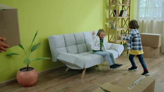In the large luminous living room two charismatic and little boys happy running to the new sofa and take a sit they enjoy the new home while parents brigs last boxes