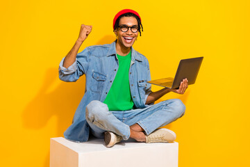Full size photo of nice young guy sitting white cube raise fist netbook wear trendy jeans outfit...