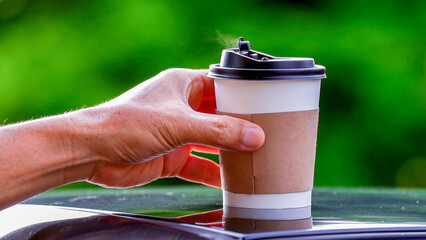 coffee takeaway in a paper cup on top of the car roof green tree background at sunrise in the morning,  selective focus, soft focus.