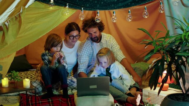 In the large extempore tent in the kids room parents and their two little boys watching movie together on the laptop while sitting inside the tent